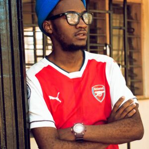 A stylish young man wearing an Arsenal jersey and blue beanie, looking thoughtful with crossed arms.